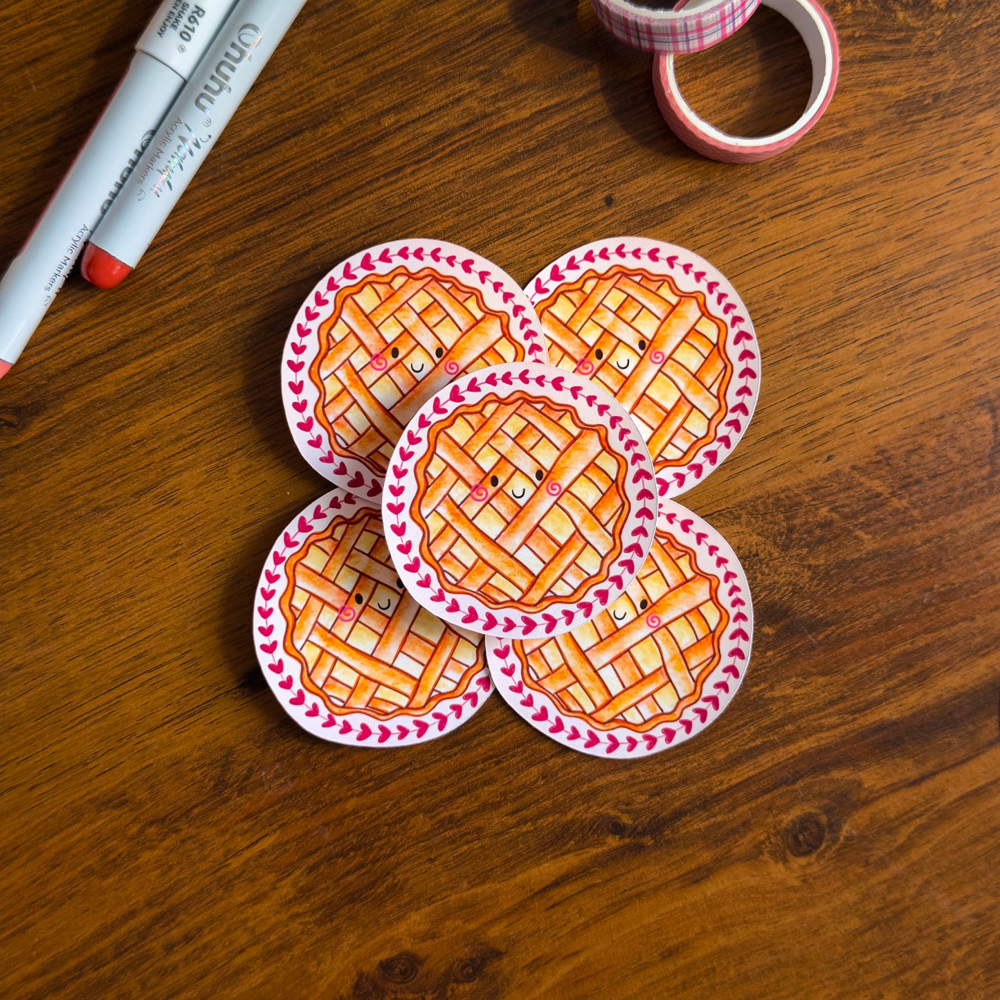 Five round stickers of a pie design on a wooden surface with markers and tape.