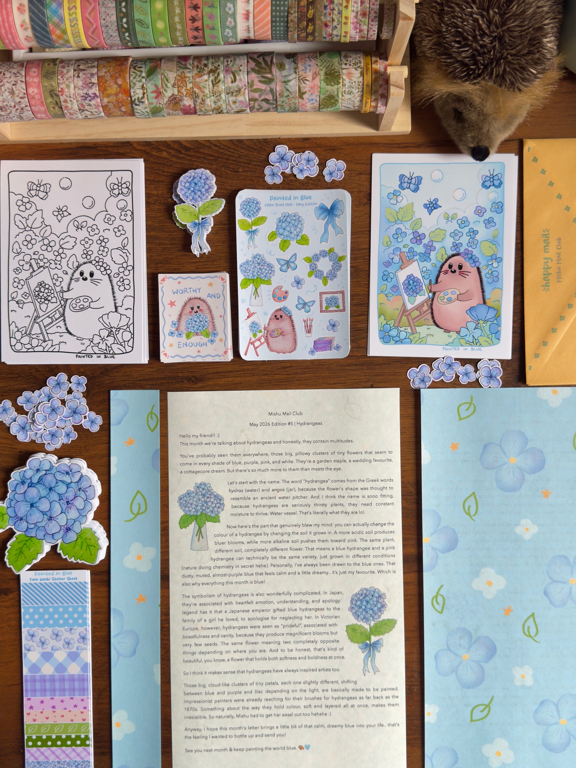 Mishu Mail Club May 2026 hydrangea edition flat lay featuring kawaii hedgehog art print, sticker sheet, die cut stickers, coloring page, themed letter, and cute envelope on wooden desk with washi tape rolls