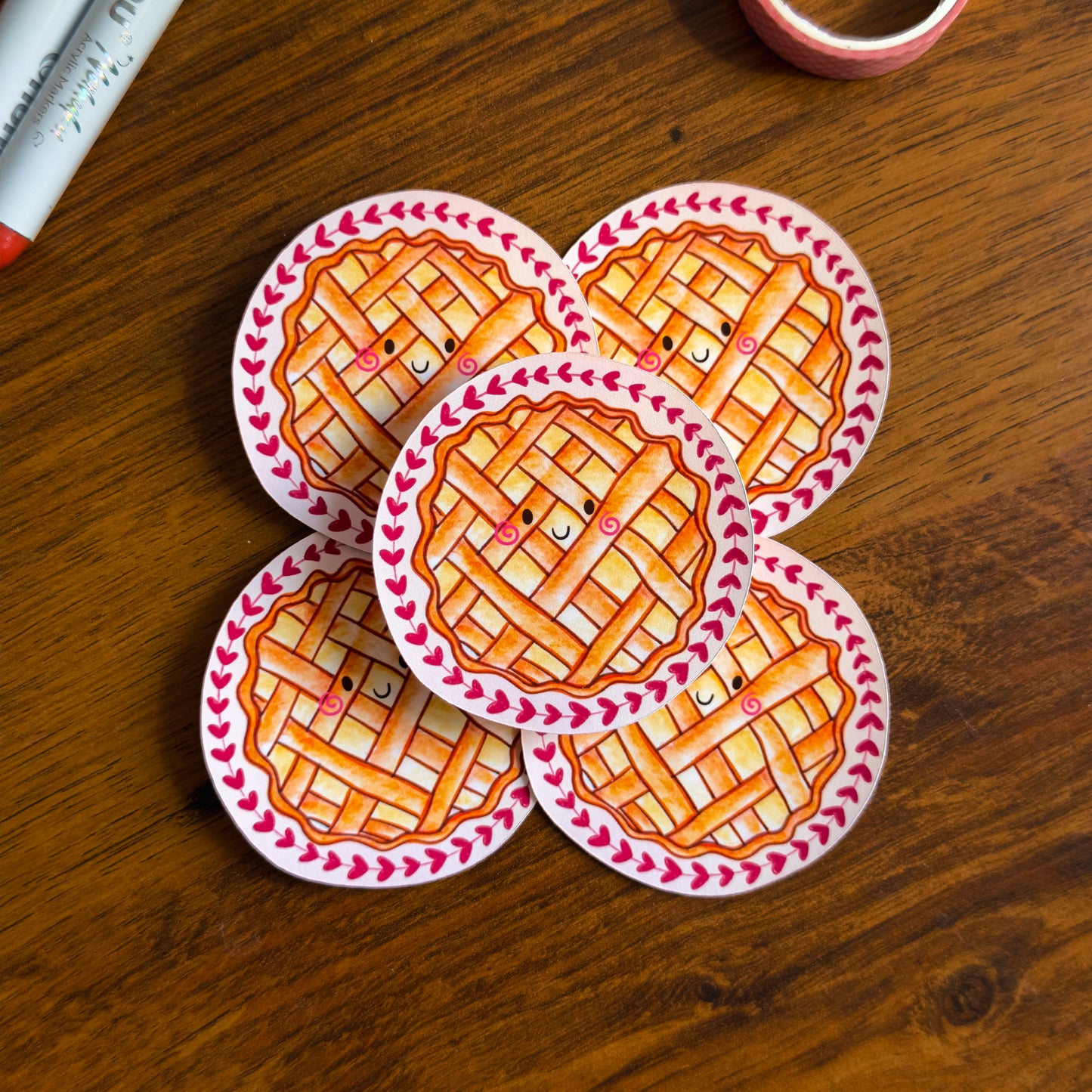 Five round stickers of a pie design on a wooden surface with markers and tape.