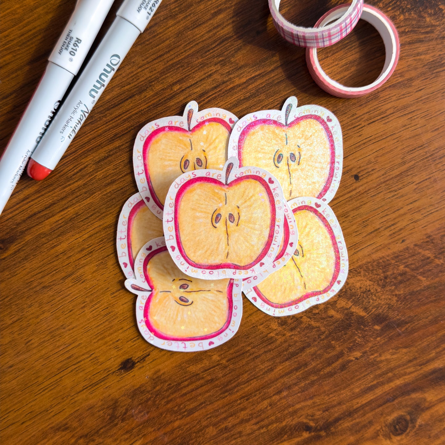 A hand-illustrated apple slice die cut sticker with holographic glitter lamination. Surrounded by the phrase "better days are coming, keep growing, keep blooming on a wooden surface