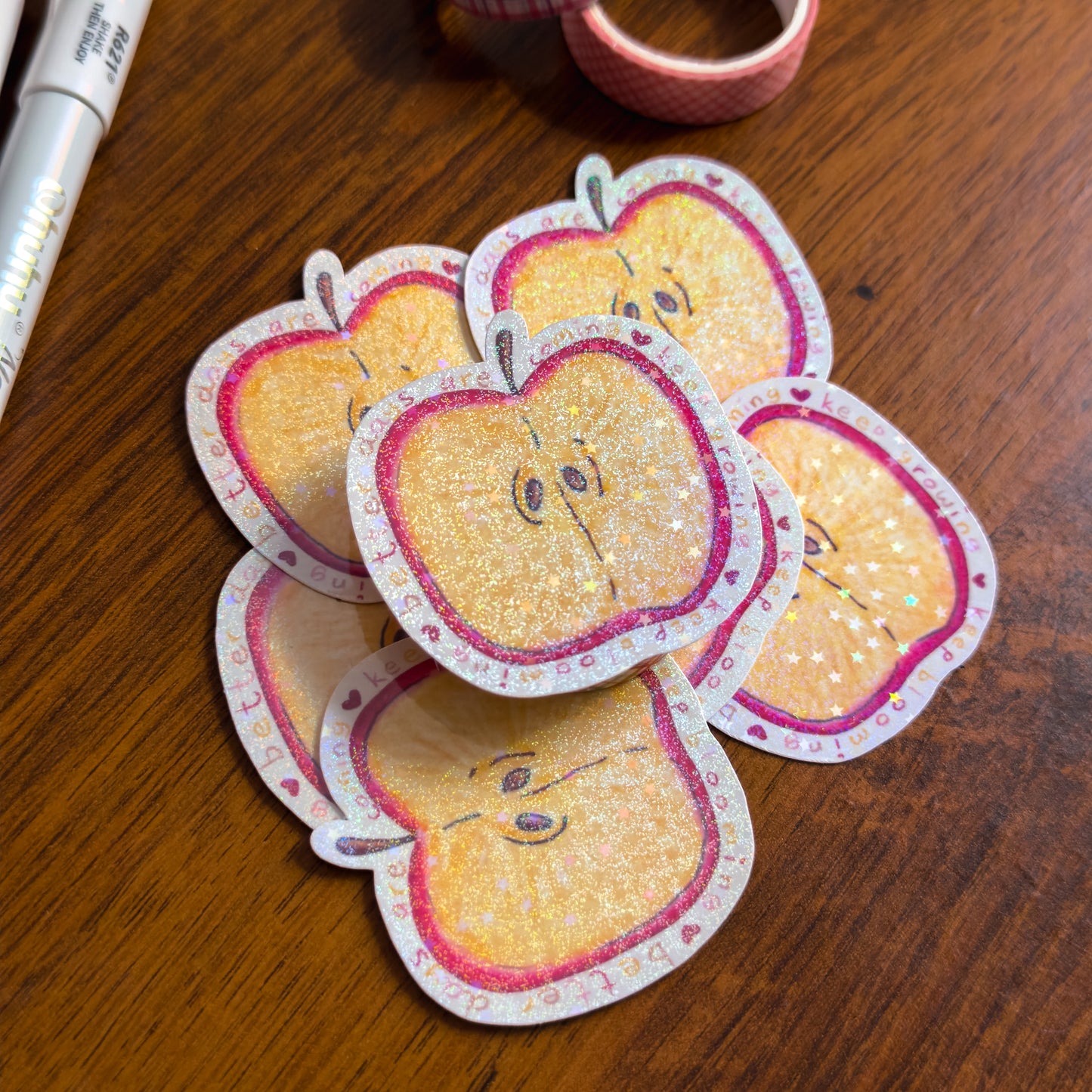 A hand-illustrated apple slice die cut sticker with holographic glitter lamination. Surrounded by the phrase "better days are coming, keep growing, keep blooming on a wooden surface
