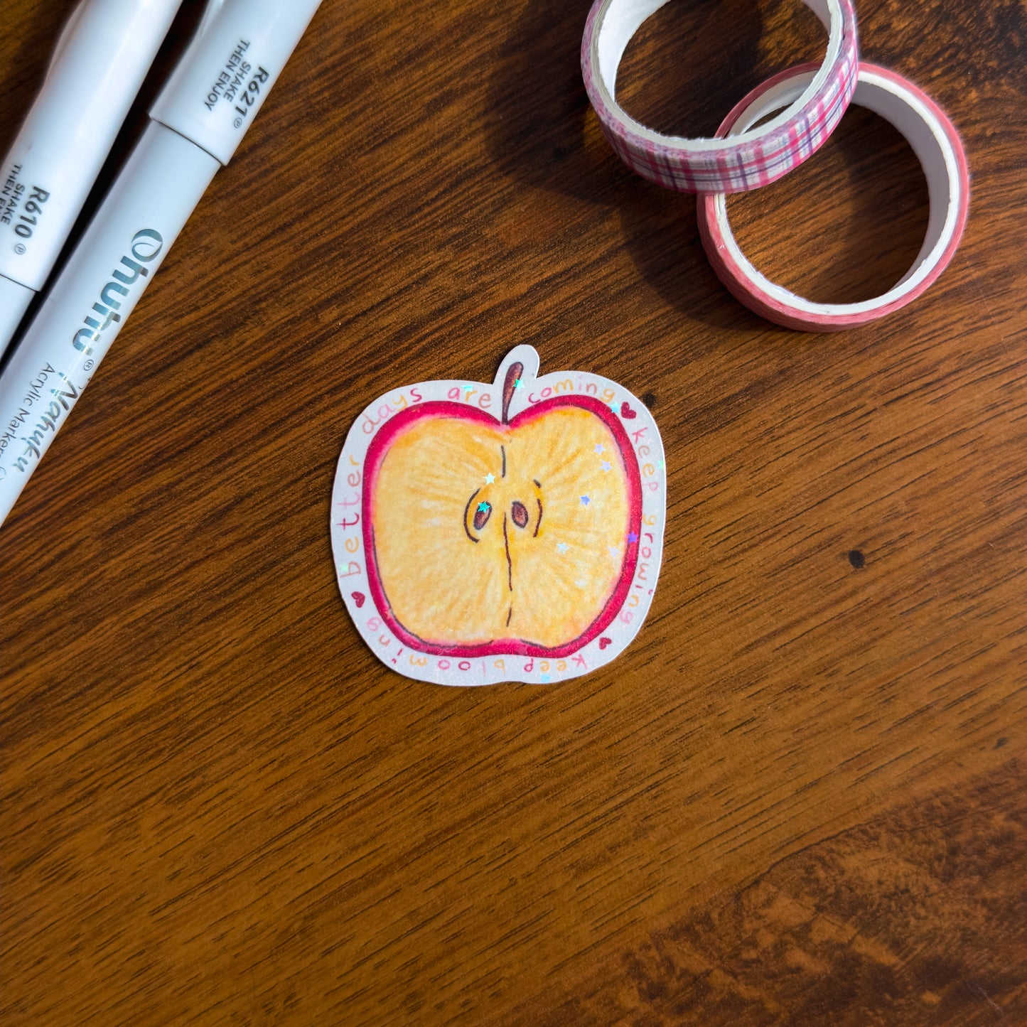 A hand-illustrated apple slice die cut sticker with holographic glitter lamination. Surrounded by the phrase "better days are coming, keep growing, keep blooming on a wooden surface