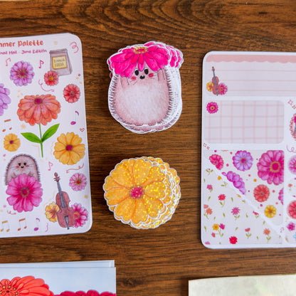 Decorative stationery items including a pink hedgehog with a flower hat, a yellow flower, and floral-patterned paper on a wooden surface.