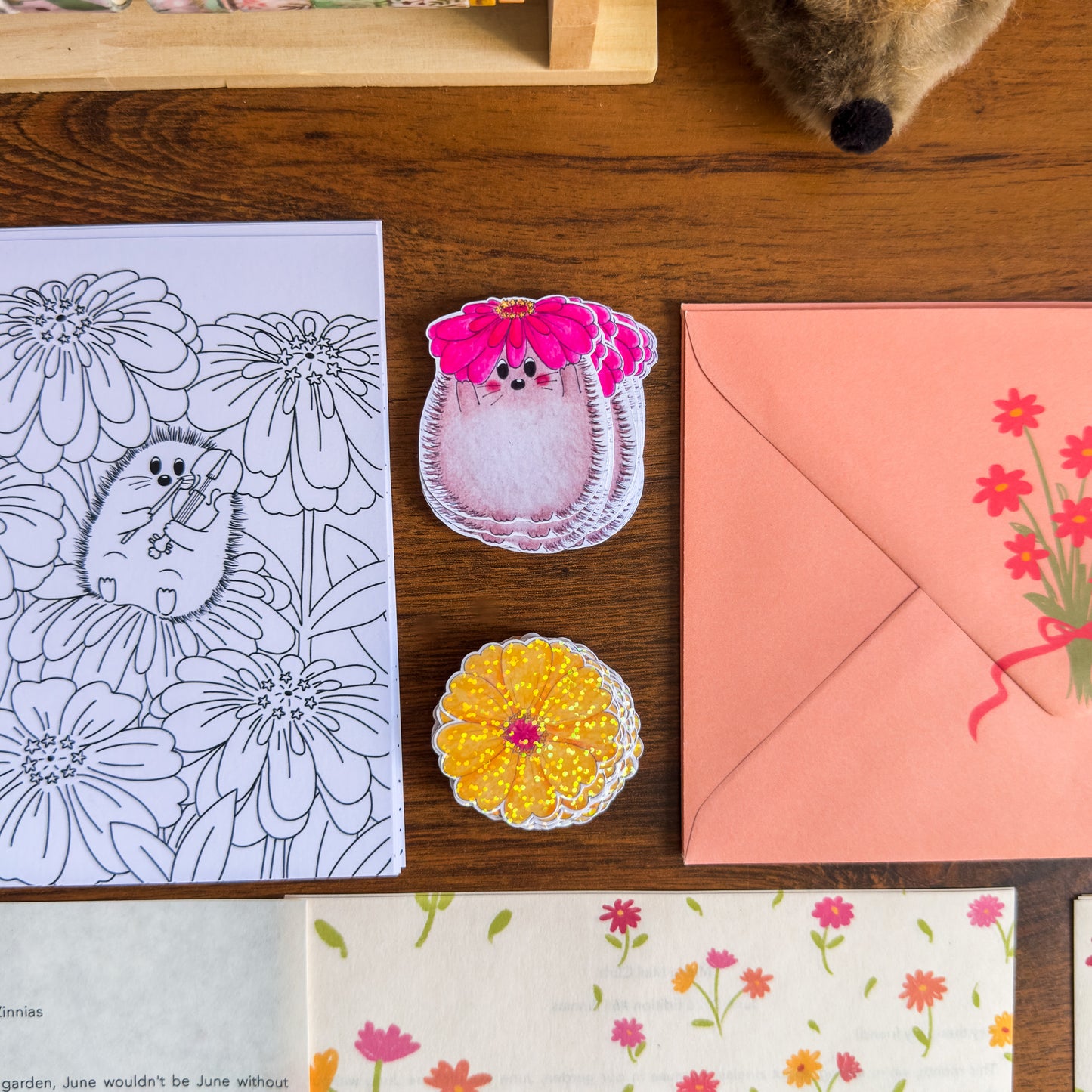 Coloring page, hedgehog holding a zinnia sticker, yellow zinnia (laminated) sticker and a pink envelope on a wooden surface