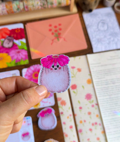 Hand holding a small hedgehog sticker holding a zinnia against a colorful background with stationery items.