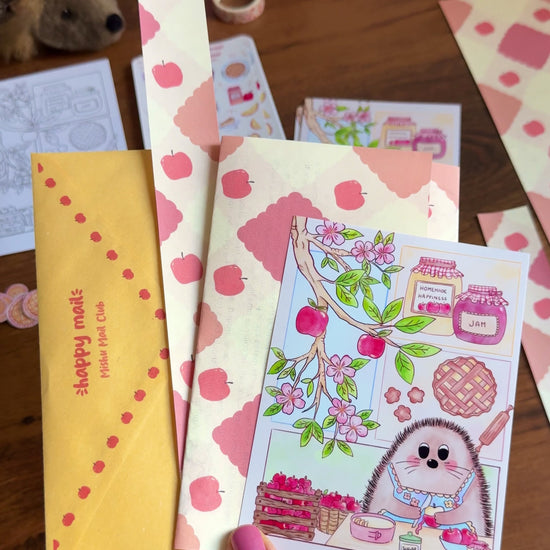 A video showing a hand holding April Mishu Mail Club contents on a wooden surface: a golden 'happy mail' envelope, apple-printed decorative paper, a 'Homemade Happiness' art print showing a hedgehog baking surrounded by apple blossoms and jam jars, and loose apple pie and apple stickers scattered nearby