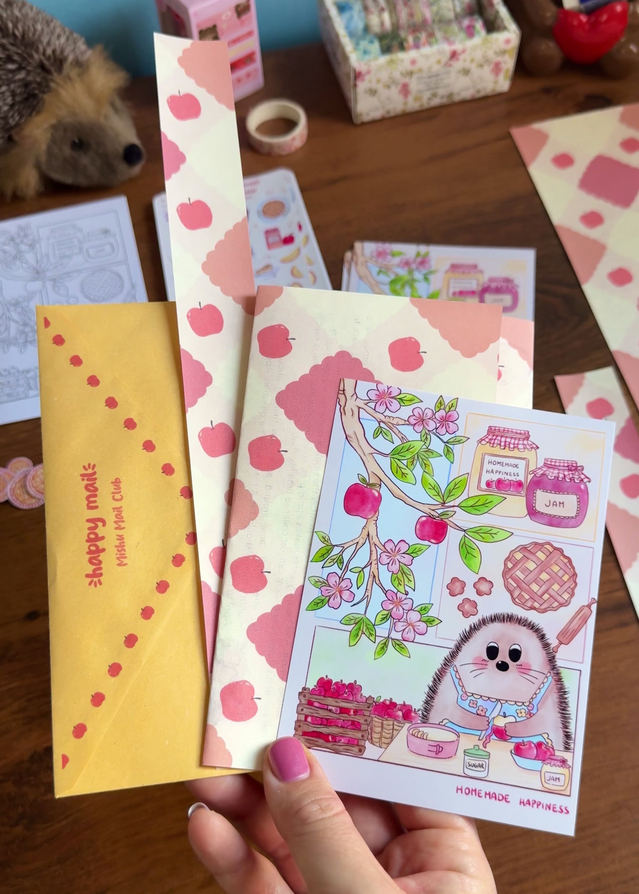A video showing a hand holding April Mishu Mail Club contents on a wooden surface: a golden 'happy mail' envelope, apple-printed decorative paper, a 'Homemade Happiness' art print showing a hedgehog baking surrounded by apple blossoms and jam jars, and loose apple pie and apple stickers scattered nearby