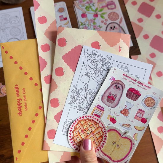 Video showing a hand holding the April Mishu Mail Club contents on a wooden surface: a yellow decorated envelope, apple-patterned paper which is the back of the letter, a 'Homemade Happiness' sticker sheet, an A6 black-and-white coloring page of the hedgehog baking scene, a kawaii apple pie matte sticker, and a holo apple slice sticker.