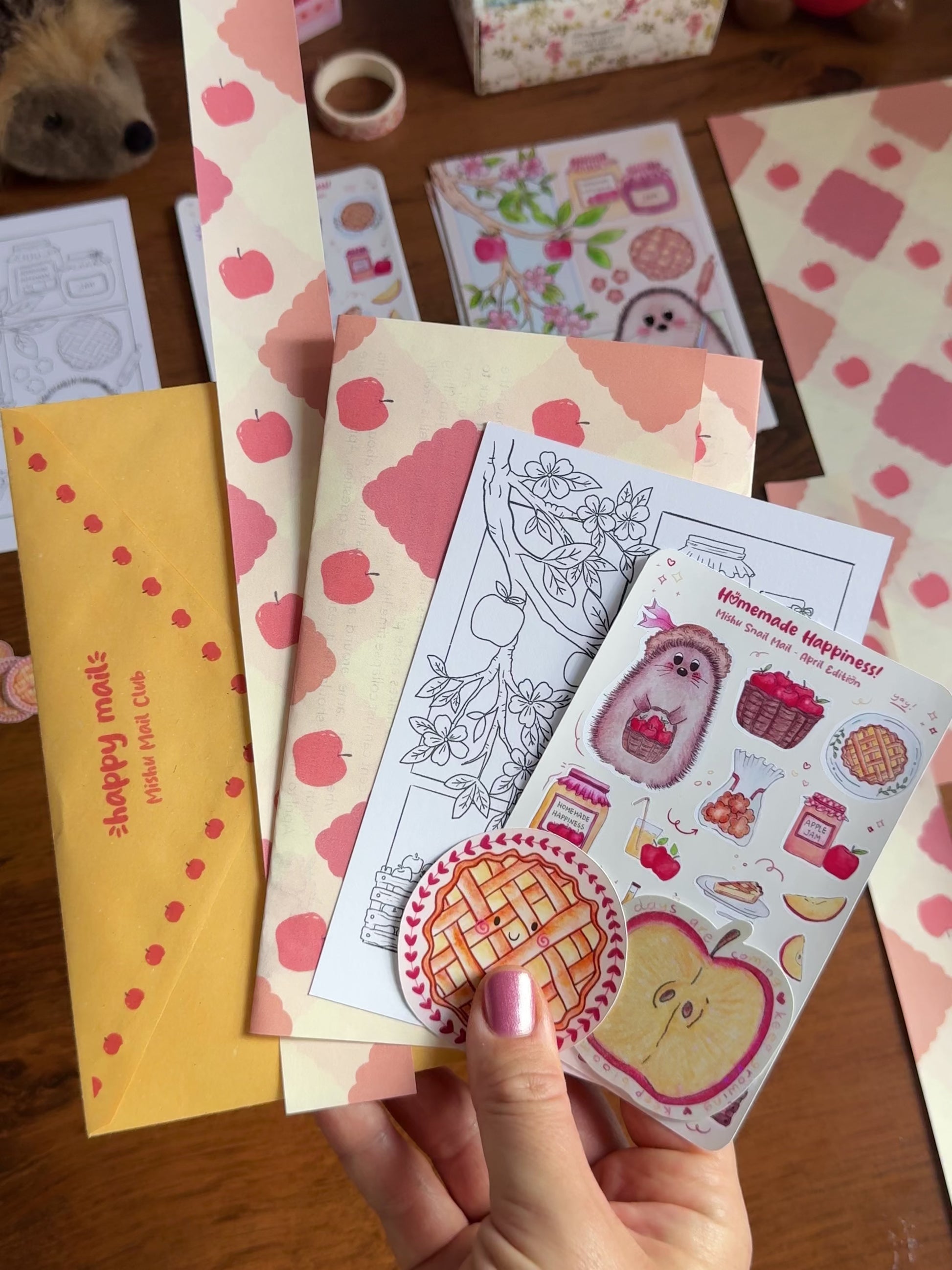 Video showing a hand holding the April Mishu Mail Club contents on a wooden surface: a yellow decorated envelope, apple-patterned paper which is the back of the letter, a 'Homemade Happiness' sticker sheet, an A6 black-and-white coloring page of the hedgehog baking scene, a kawaii apple pie matte sticker, and a holo apple slice sticker.