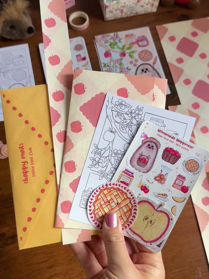 Video showing a hand holding the April Mishu Mail Club contents on a wooden surface: a yellow decorated envelope, apple-patterned paper which is the back of the letter, a 'Homemade Happiness' sticker sheet, an A6 black-and-white coloring page of the hedgehog baking scene, a kawaii apple pie matte sticker, and a holo apple slice sticker.