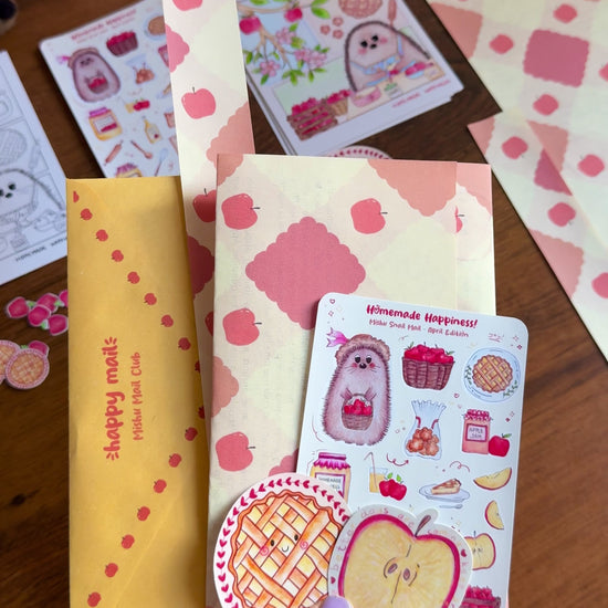 Video showing a hand holding the April Mishu Mail Club contents on a wooden surface: a golden envelope, apple-patterned paper, a 'Homemade Happiness' sticker sheet, a round kawaii apple pie matte sticker, and a holo glittery apple slice sticker, with extra stickers scattered around