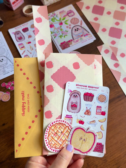 Video showing a hand holding the April Mishu Mail Club contents on a wooden surface: a golden envelope, apple-patterned paper, a 'Homemade Happiness' sticker sheet, a round kawaii apple pie matte sticker, and a holo glittery apple slice sticker, with extra stickers scattered around
